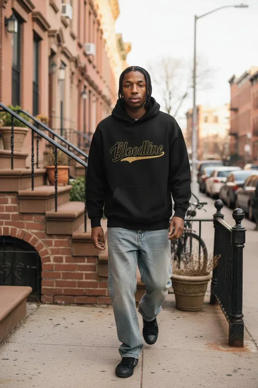 A man walking on a city sidewalk wearing a black hoodie with a gold 'Vindoline' graphic across the chest, paired with light blue jeans and black shoes.