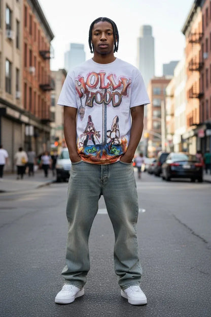 A man stands in the middle of a city street wearing a white graphic t-shirt with a colorful 'Hollywood' print, light gray jeans, and white sneakers.