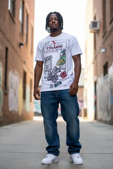 A man wearing a white graphic t-shirt with 'HOOD MAGAZINE' printed on the front, paired with blue jeans and white sneakers, standing in an urban alleyway.
