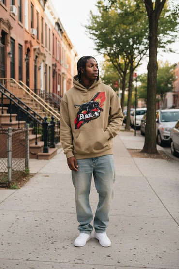A man stands on a city sidewalk wearing a beige hoodie with a black and red graphic that reads 'BLOODYLINE', paired with light blue jeans and white sneakers.