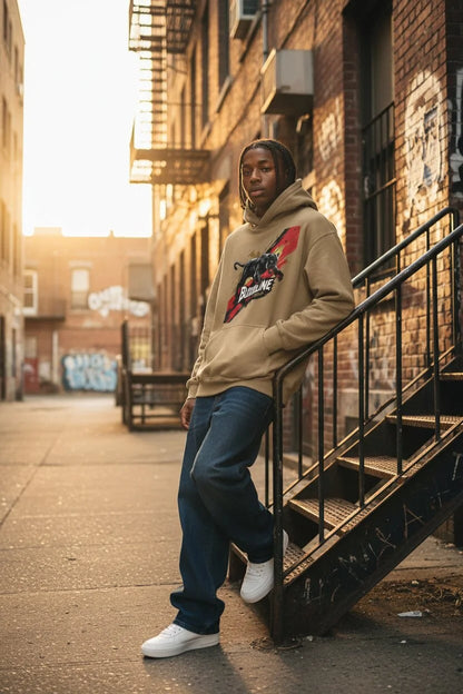A person standing on an outdoor stairway in an urban alley, wearing a beige hoodie with a red and black graphic print, dark blue jeans, and white sneakers.