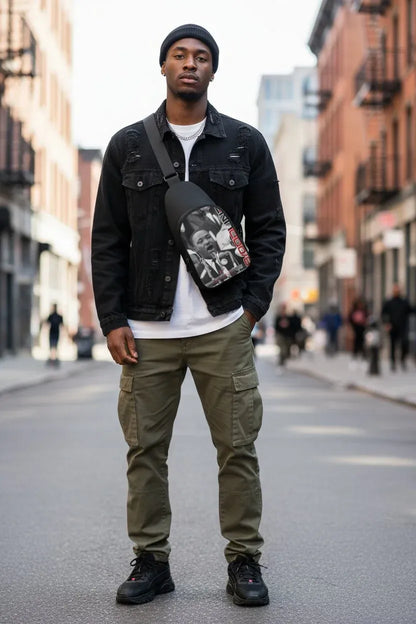 A man stands on a city street wearing a black denim jacket, black beanie, white t-shirt, olive green cargo pants, and black sneakers, with a black crossbody bag featuring a graphic print across his chest.