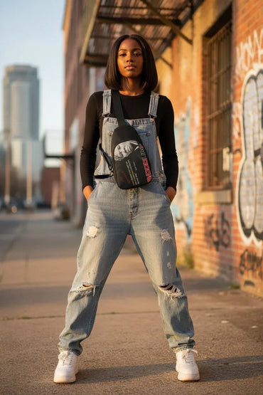 A woman stands on a city sidewalk wearing light-wash distressed denim overalls over a black long-sleeve top, with a black crossbody bag featuring a graphic print and pink text. The background shows a brick wall with graffiti and a blurred urban street scene.