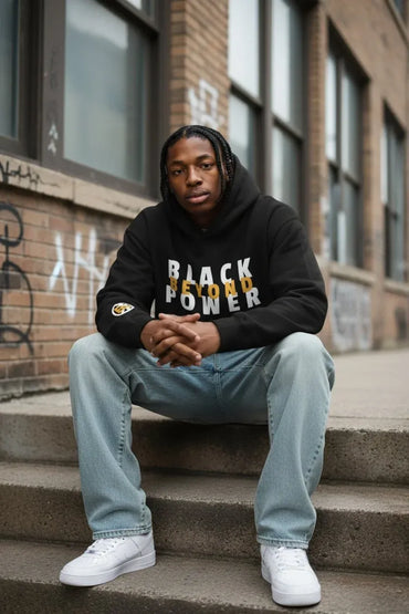 A man sitting on concrete steps in front of a brick building, wearing a black hoodie with 'BLACK POWER' printed in white and gold, light blue jeans, and white sneakers.