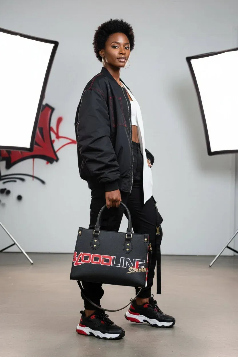 A model stands in a studio setting holding a black tote bag with 'VOODLINE' printed in red and white on the front. The model is wearing a black bomber jacket, white shirt, black pants, and black sneakers with red accents.
