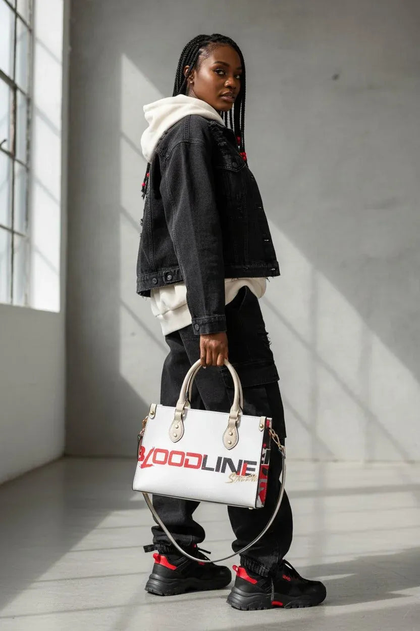 A model stands in a minimalist concrete room holding a white handbag with 'GOODLINE' printed in red and black. The model is wearing a black denim jacket, white hoodie, black cargo pants, and black sneakers with red accents.