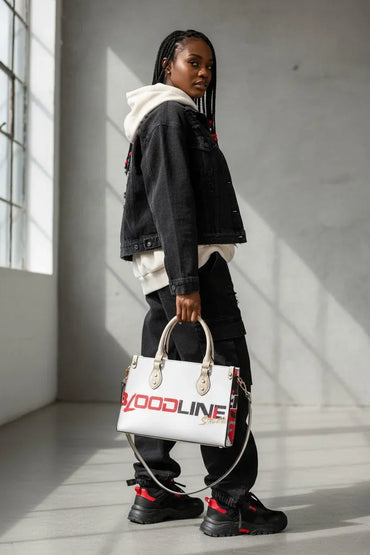 A model stands in a minimalist concrete room holding a white handbag with 'GOODLINE' printed in red and black. The model is wearing a black denim jacket, white hoodie, black cargo pants, and black sneakers with red accents.