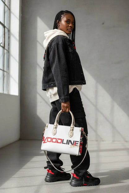 A model stands in a minimalist concrete room holding a white handbag with 'GOODLINE' printed in red and black. The model is wearing a black denim jacket, white hoodie, black cargo pants, and black sneakers with red accents.