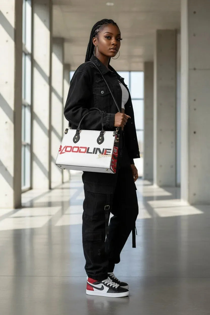 A model stands in a modern concrete hallway carrying a white 'GOODLINE' tote bag with red and black accents. The model is dressed in a black denim jacket, black cargo pants, and black and white sneakers.