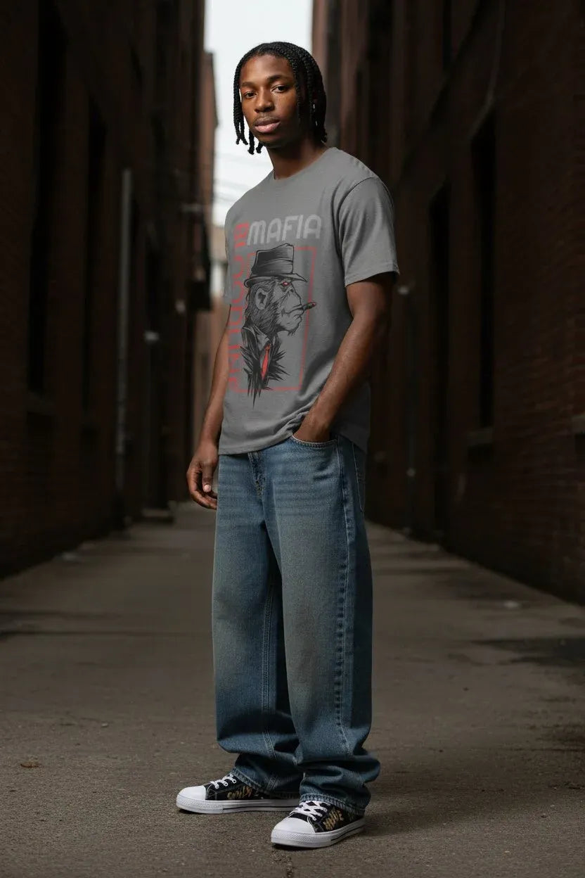 A man wearing a gray graphic t-shirt with a red and black design, blue jeans, and white sneakers, standing in a narrow brick alleyway.