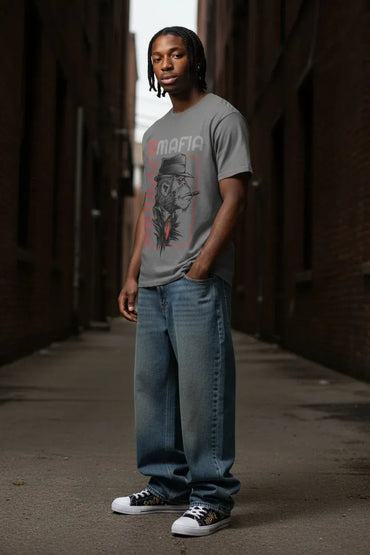 A man wearing a gray graphic t-shirt with a red and black design, blue jeans, and white sneakers, standing in a narrow brick alleyway.