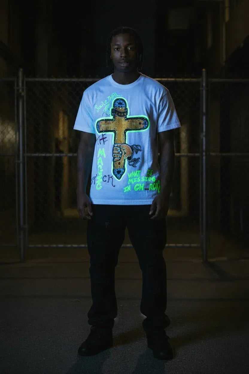 A man wearing a light blue graphic t-shirt with a large cross design and neon green text, standing in a dark alley at night.