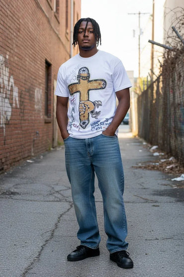 A man standing in an urban alleyway wearing a white graphic t-shirt with a brown cross design, blue jeans, and black shoes.