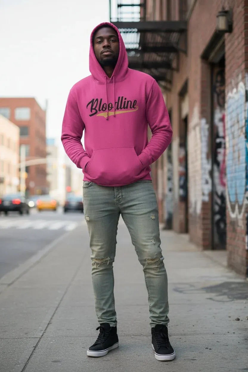 A man standing on a city sidewalk wearing a bright pink hoodie with 'Bloodline' printed across the chest in black script with a gold underline, paired with light green distressed jeans and black sneakers.