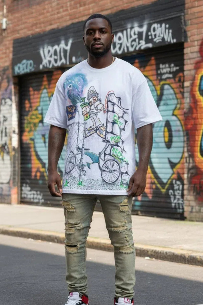 A man stands on a city street wearing a white graphic t-shirt with a bicycle illustration, olive green distressed jeans, and white, red, and black sneakers. The background features a brick wall and graffiti-covered metal garage doors.