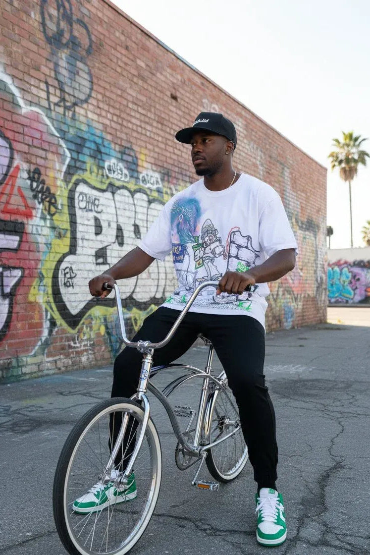 A man wearing a white graphic t-shirt, black pants, and green and white sneakers sits on a silver cruiser bicycle in front of a graffiti-covered brick wall.