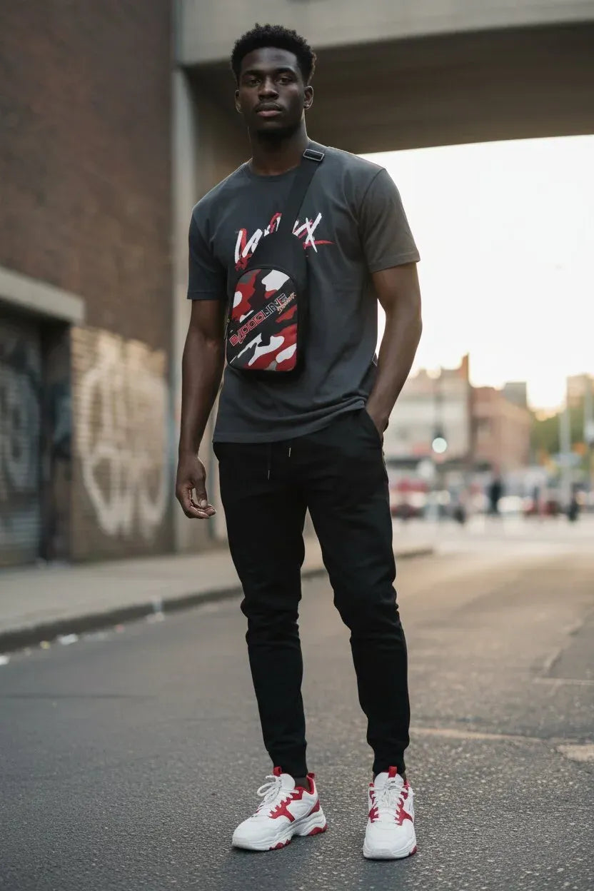 A man standing on an urban street under a bridge, wearing a dark gray graphic t-shirt, black jogger pants, and white sneakers with red accents.