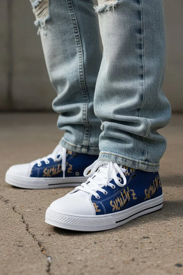 A close-up of a person's legs wearing light-wash distressed jeans and blue low-top sneakers with white laces, white rubber toe caps and soles, and gold graffiti-style lettering on the sides, standing on concrete.
