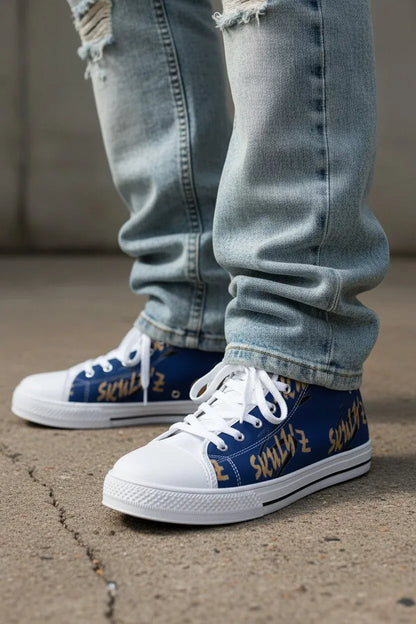 A close-up of a person's legs wearing light-wash distressed jeans and blue low-top sneakers with white laces, white rubber toe caps and soles, and gold graffiti-style lettering on the sides, standing on concrete.