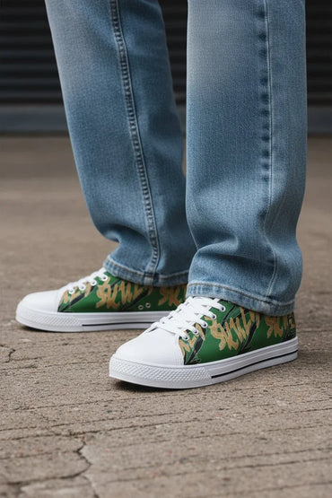 A close-up of a person's feet wearing green and tan camouflage low-top sneakers with white rubber soles, paired with light-wash blue jeans, standing on a concrete surface.