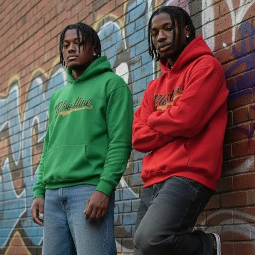 Two men standing against a graffiti-covered brick wall, wearing matching hoodie styles in green and red with blue jeans and black sneakers.