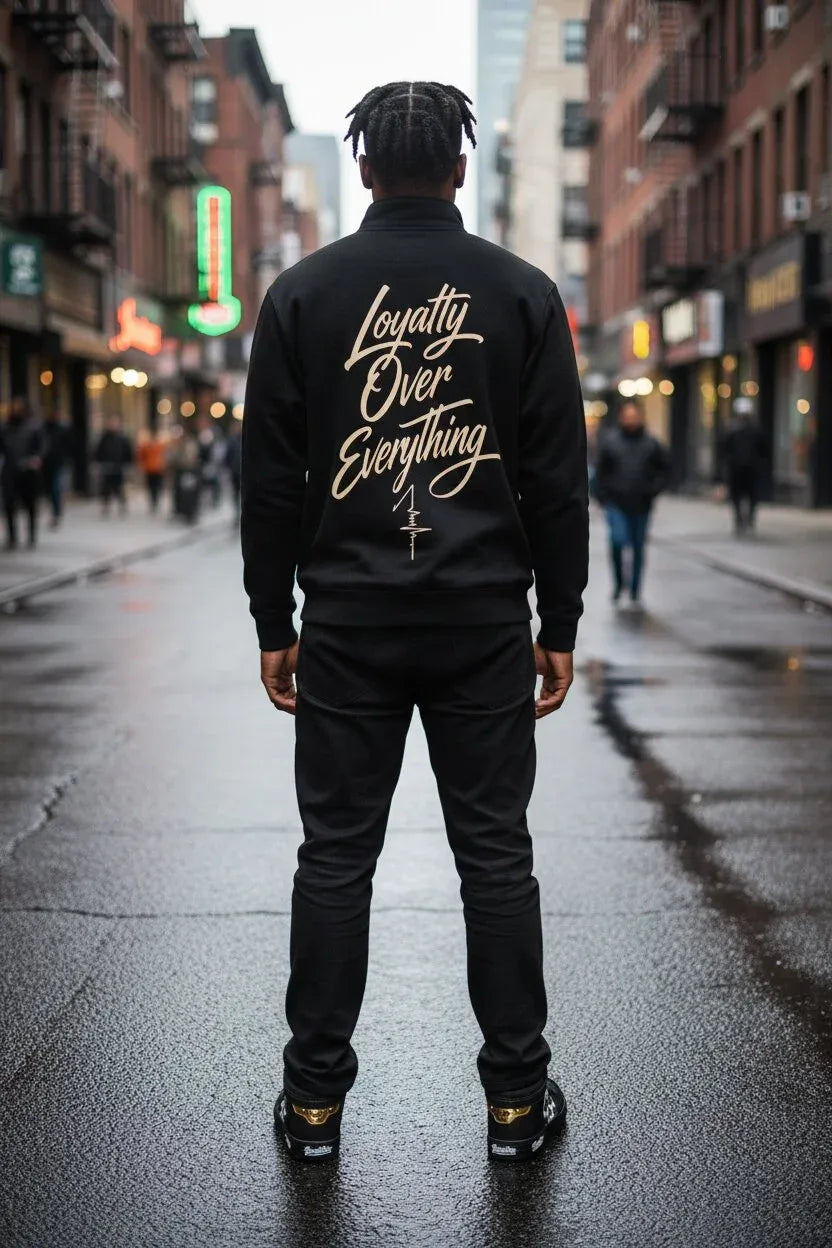 A man stands on a wet city street facing away from the camera, wearing a black bomber jacket with 'Loyalty Over Everything' printed in gold script across the back, paired with black pants and sneakers.