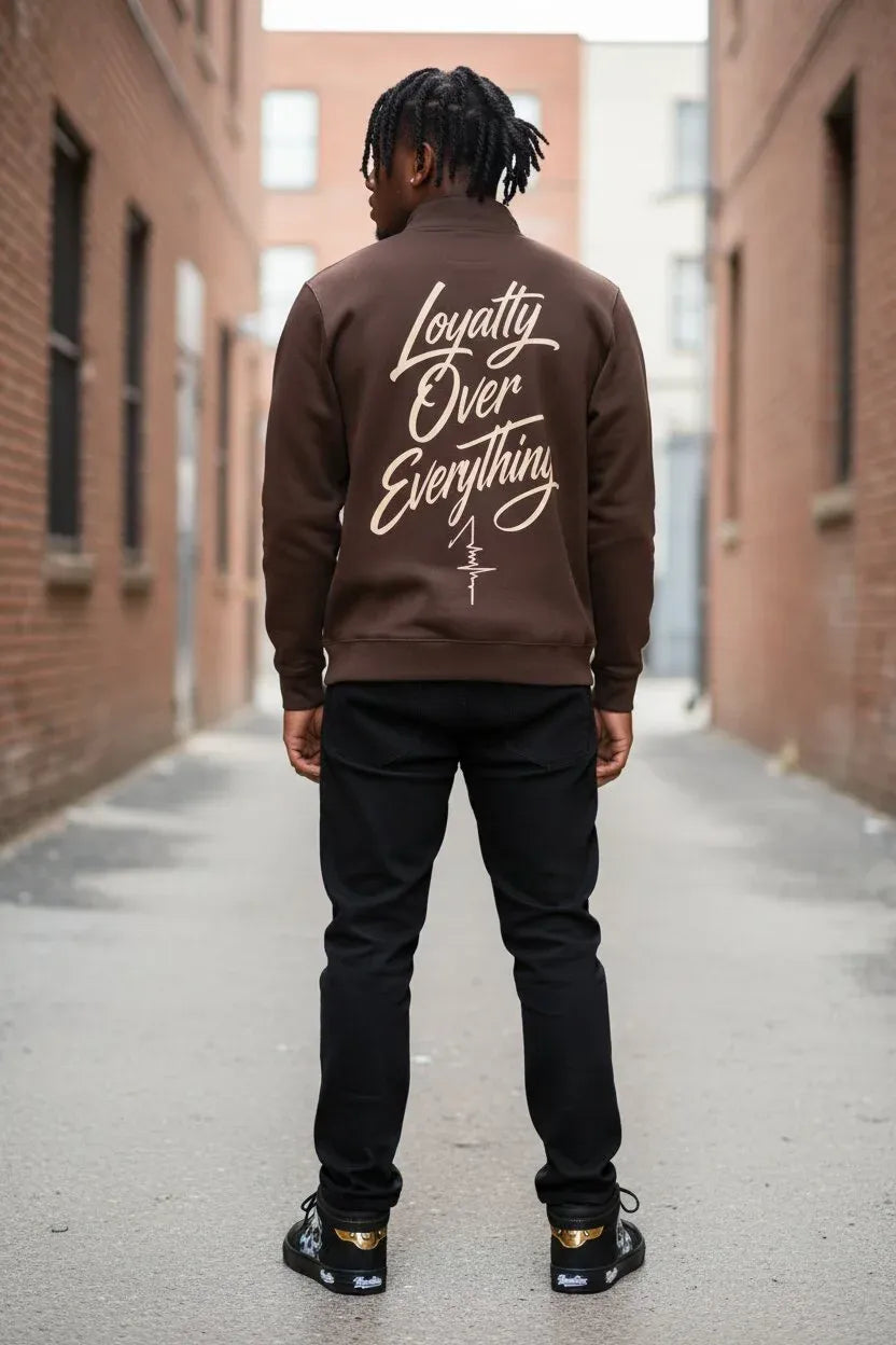 A man viewed from behind wearing a brown hoodie with 'Loyalty Over Everything' printed in white script on the back, paired with black pants and black sneakers, standing in an alleyway between brick buildings.
