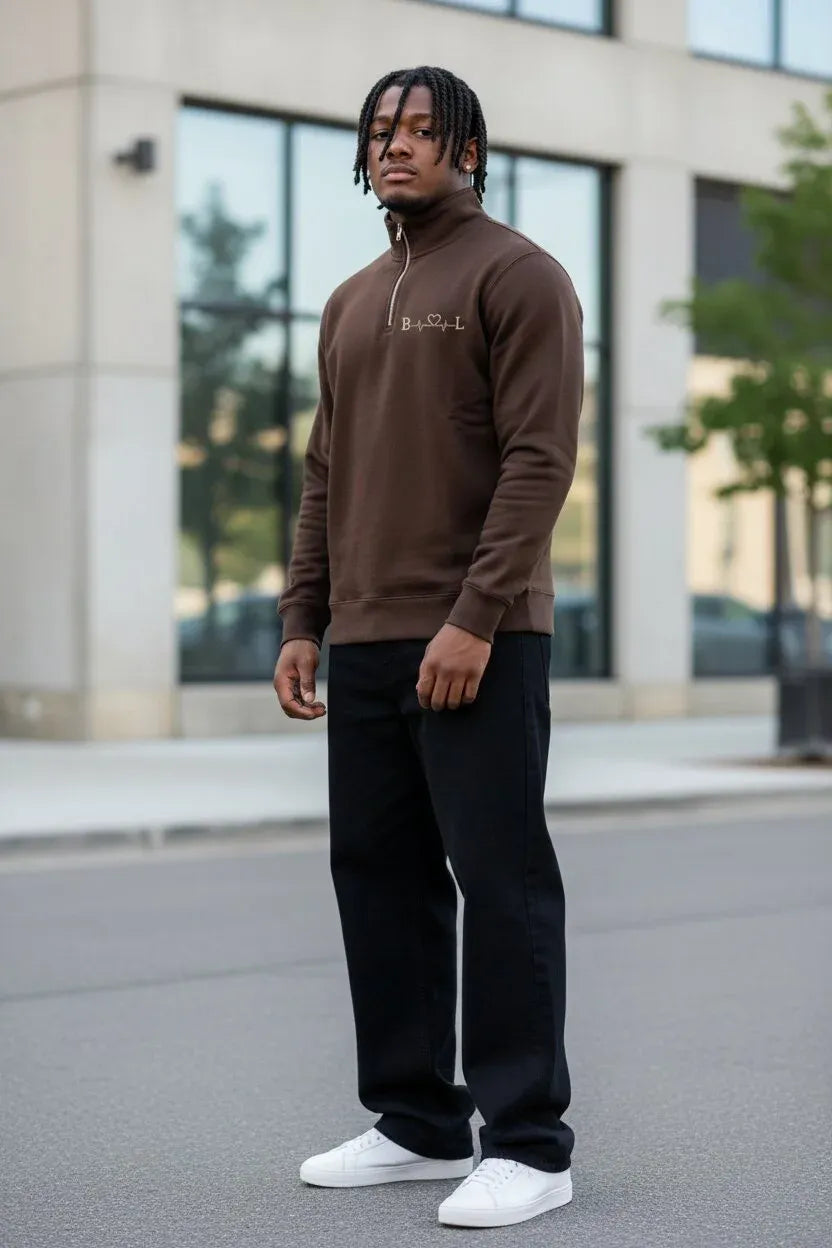 A man wearing a brown quarter-zip sweatshirt with a small gold embroidered logo on the chest, paired with black pants and white sneakers, standing outdoors in front of a modern building.