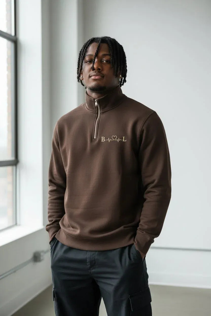 A man wearing a brown quarter-zip sweatshirt with a small embroidered logo on the chest, paired with dark cargo pants, standing in a bright indoor setting.