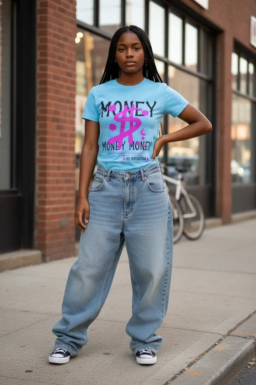 A woman stands on a city sidewalk wearing a light blue graphic T-shirt with pink and black text, light-wash wide-leg jeans, and black-and-white sneakers. The background shows a brick building, storefront windows, and a bicycle.