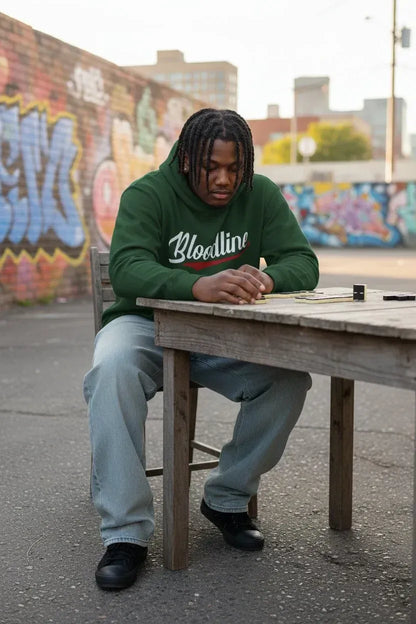 A man wearing a green hoodie with 'Blocelline' written on the front, sitting at a wooden table outdoors against a graffiti-covered wall.