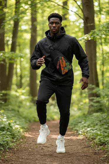 A man jogging on a forest path wearing a black hooded jacket, black sweatpants, and white sneakers, with a black crossbody bag featuring a green and orange graphic design.