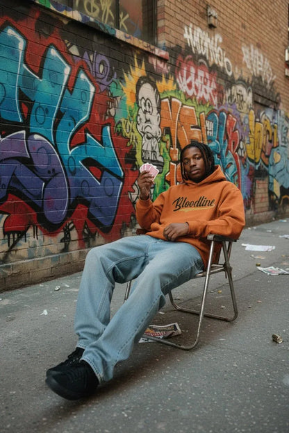 A man sits on a metal folding chair in front of a graffiti-covered brick wall, wearing an orange hoodie with 'Bloodline' printed across the chest, light blue jeans, and black sneakers.