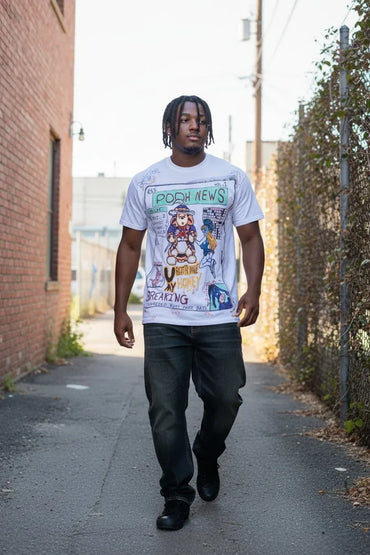 A man walking down an outdoor alley wearing a white graphic t-shirt with 'Pooh News' printed on the front, paired with dark gray jeans and black shoes.