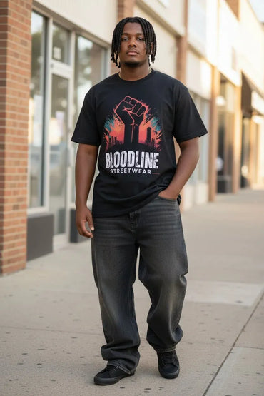A man standing on an outdoor city sidewalk wearing a black graphic t-shirt with a raised fist design and the text 'BLOODLINE STREETWEAR', paired with dark gray jeans and black sneakers.