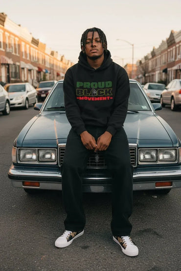 A man wearing a black hoodie with a green and red 'PROUD BLACK MOVEMENT' graphic sits on the front bumper of a dark blue classic car on a city street.