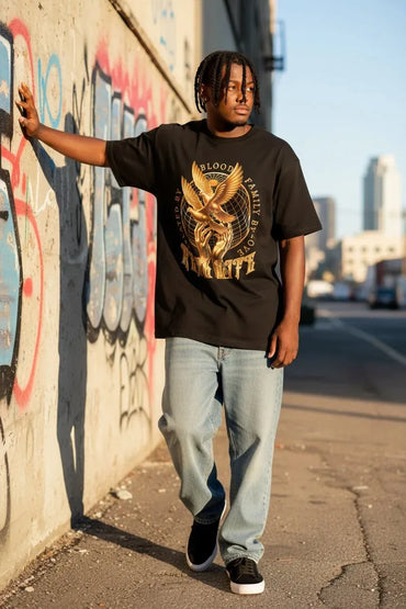 A man wearing a black graphic t-shirt with a gold eagle design and the words 'Blood Family' and 'Anchorage', paired with light blue jeans and black sneakers, standing on a city sidewalk next to a graffiti-covered wall.