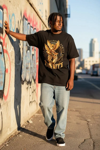 A man wearing a black graphic t-shirt with a gold eagle design and the words 'Blood Family' and 'Anchorage', paired with light blue jeans and black sneakers, standing on a city sidewalk next to a graffiti-covered wall.