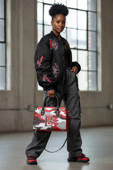 A model stands in a loft-style room with large windows, wearing a black bomber jacket with red and white embroidery, dark gray cargo pants, and red sneakers, while holding a red, white, and gray camouflage handbag.