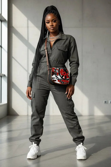 A woman modeling a dark gray utility jumpsuit with a red, black, and white patterned crossbody bag, styled with white sneakers in a modern concrete room.