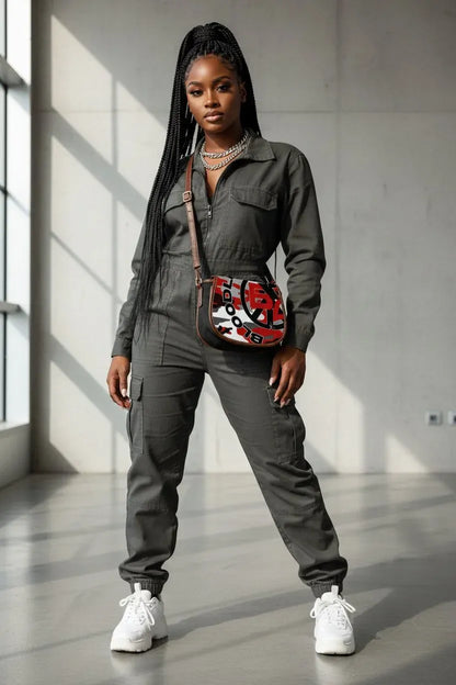 A woman modeling a dark gray utility jumpsuit with a red, black, and white patterned crossbody bag, styled with white sneakers in a modern concrete room.