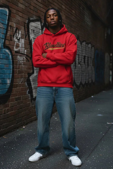 A man wearing a bright red hoodie with 'Bloodyline' written in black script across the chest, paired with blue jeans and white sneakers, standing in front of a graffiti-covered brick wall.