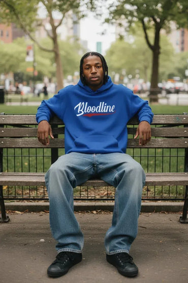 A man sitting on a wooden park bench wearing a bright blue hoodie with 'Bloodline' printed in white script across the chest, paired with light blue jeans and black sneakers.