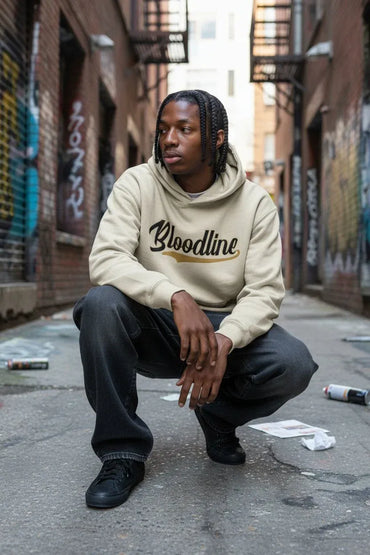 A man crouching in an urban alley wearing a beige Bloodline hoodie, black sweatpants, and black sneakers.