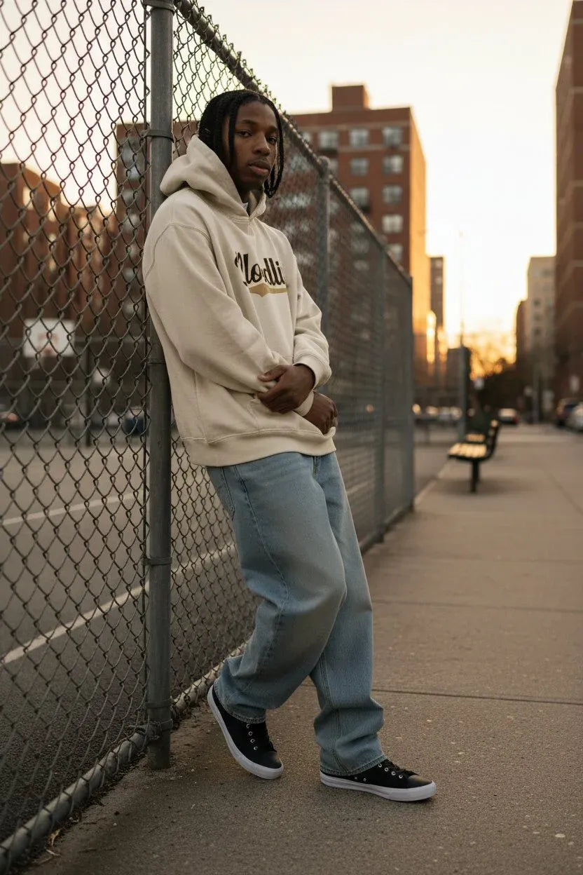 A man wearing a cream-colored hoodie with a gold script logo, light blue jeans, and black sneakers leans against a chain-link fence on a city sidewalk during sunset.