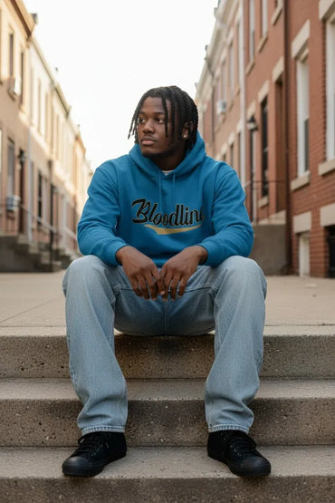 A man sitting on concrete steps in an urban street setting, wearing a bright blue hoodie with 'Bloodline' printed across the chest in black script, paired with light blue jeans and black sneakers.