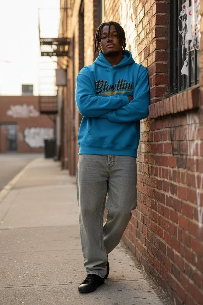 A man stands on a city sidewalk against a brick wall, wearing a bright blue hoodie with 'Bloodline' printed across the chest in black and gold, paired with light gray jeans and black shoes.