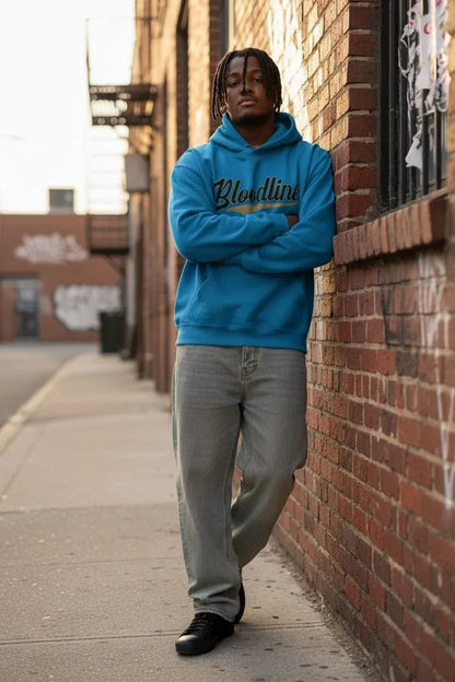A man stands on a city sidewalk against a brick wall, wearing a bright blue hoodie with 'Bloodline' printed across the chest in black and gold, paired with light gray jeans and black shoes.
