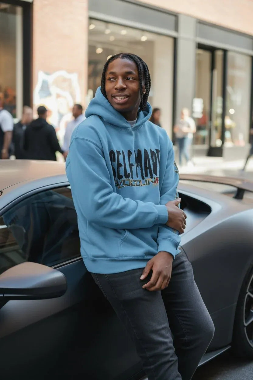 A man wearing a light blue pullover hoodie with 'SELF MADE' printed in black across the chest, leaning against a dark gray sports car on a city street.