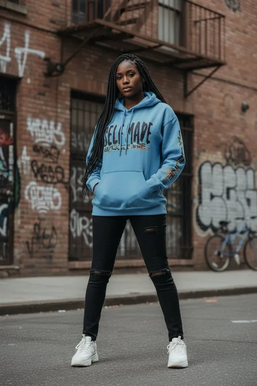 A woman stands on a city street wearing a light blue hoodie with 'SELF MADE' printed across the chest, black ripped jeans, and white sneakers, with a graffiti-covered brick building in the background.